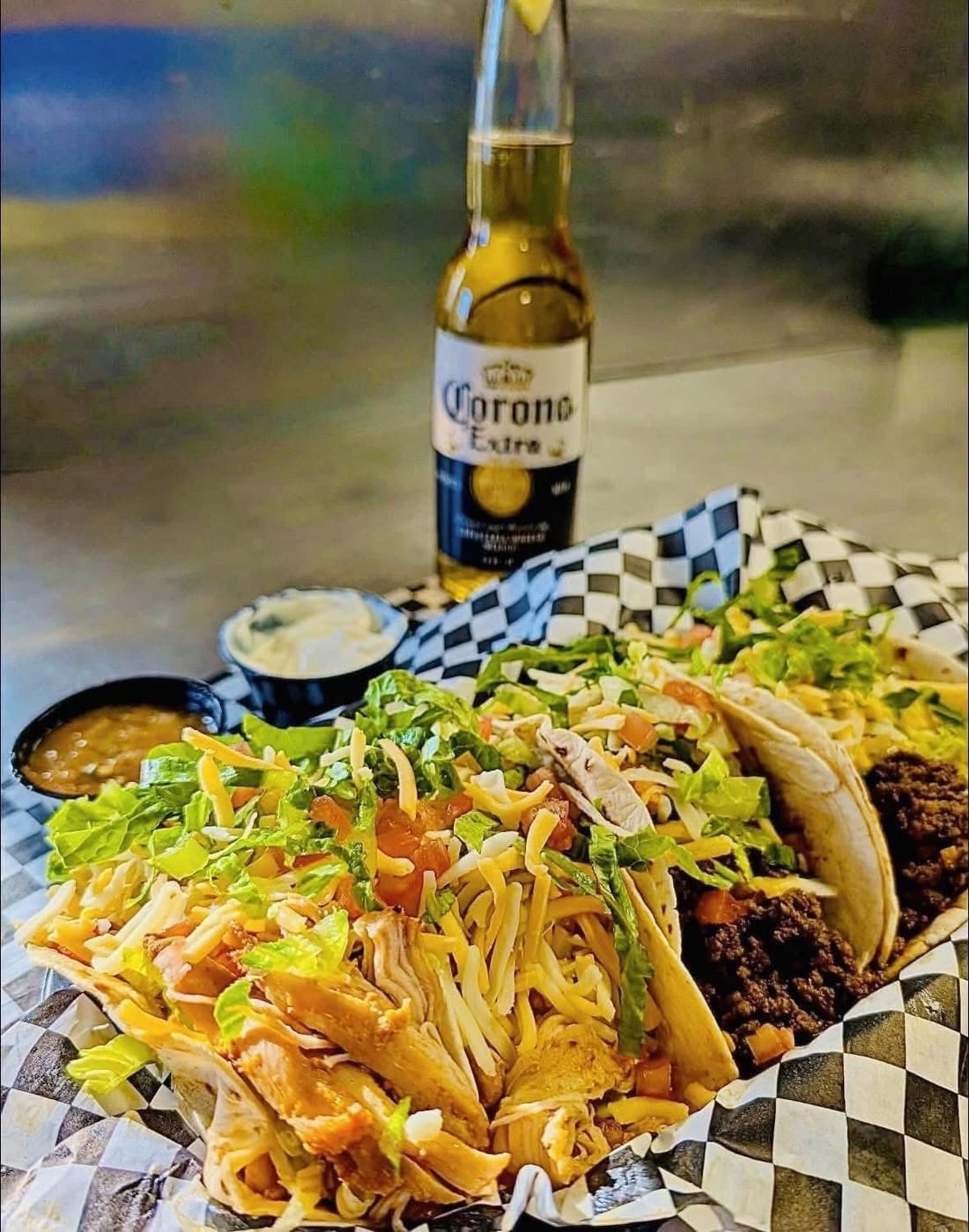 Tacos loaded with lettuce and cheese in a checkered basket, with a Corona